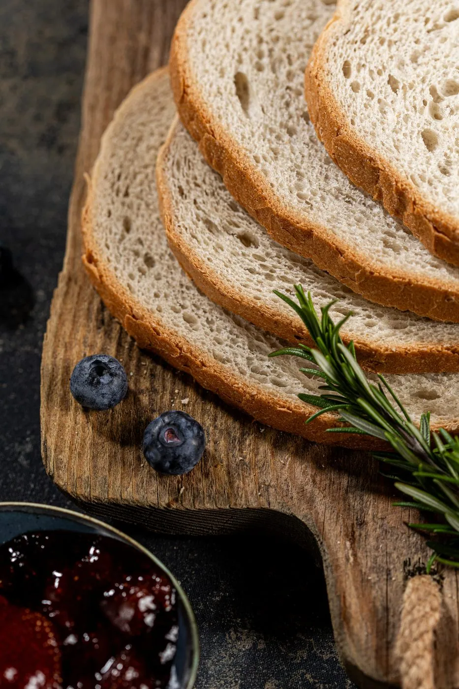 Sliced artisan rye bread on a wooden board with blueberry preserves and rosemary
